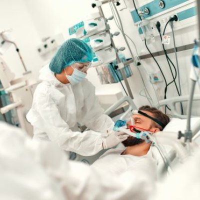 A person in a medical gown, mask and hair covering bends over a man in a bed surrounded by medical equipment.